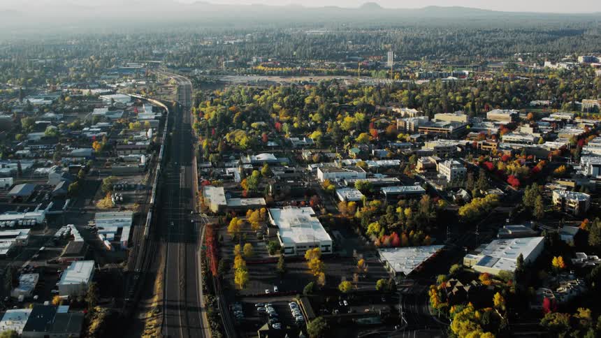 An aerial view of a city showcasing a mix of residential and commercial areas nestled amidst lush green trees and a body of water with distant mountains adding to the scenic backdrop