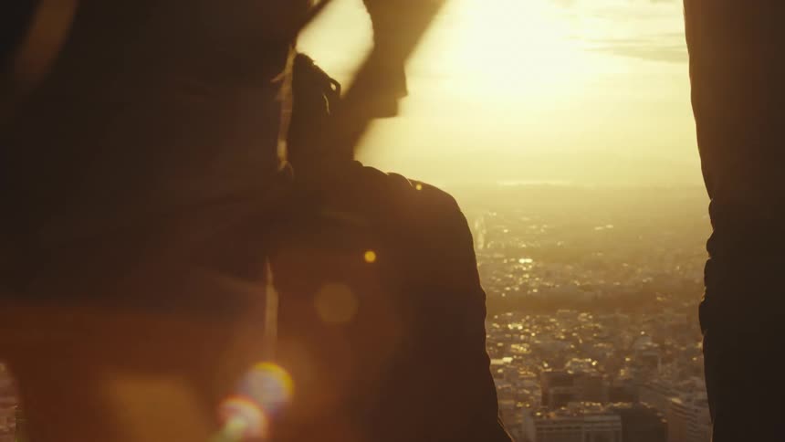 A person with dreadlocks is silhouetted against a golden sunset overlooking a sprawling cityscape below with lens flares adding warmth to the atmospheric scene