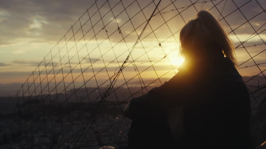 A silhouette of a person with a ponytail sits pensively by a wire fence gazing at a serene sunset over a cityscape