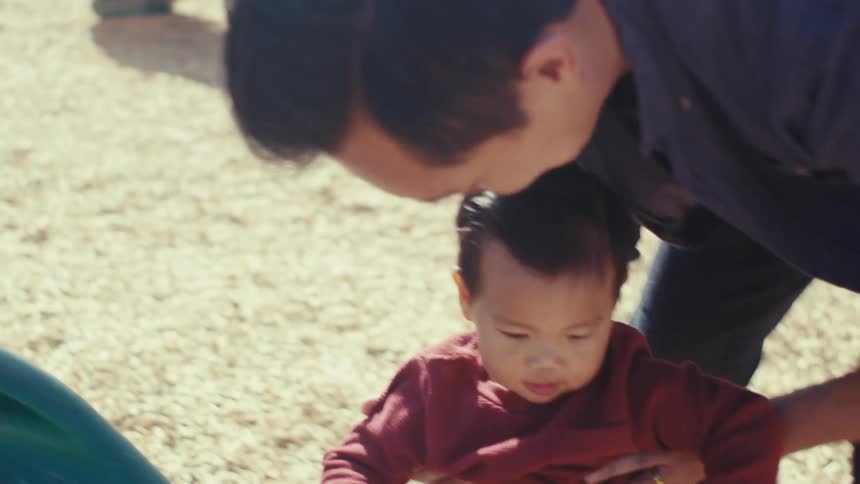 Boy going down a slide at a playground and his father catching him and wiping his chest