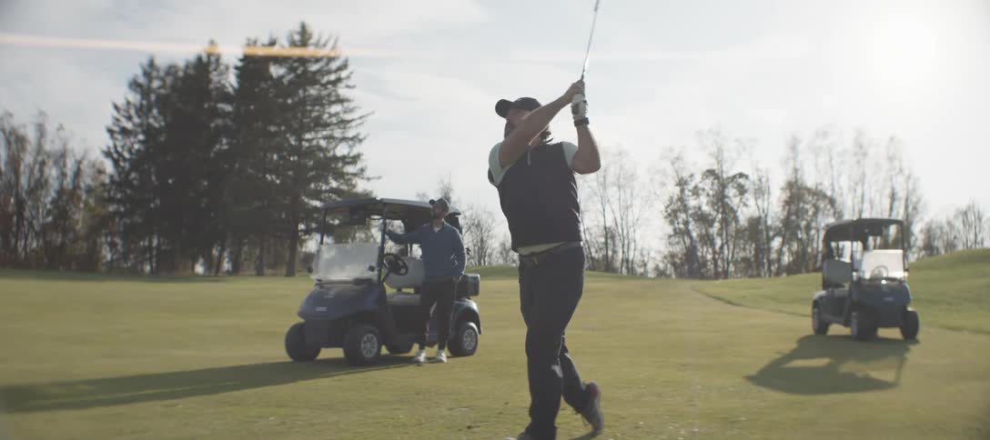 A golfer is striking the ball on a lush golf course while another person stands near golf carts watching surrounded by open greenery and trees under a calm cloudy sky