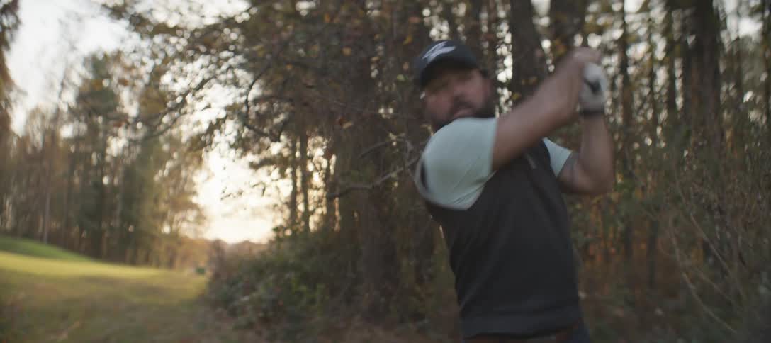 A golfer midswing wearing a dark vest light shirt and cap set against a serene wooded backdrop with tall trees and soft natural light filtering through the branches