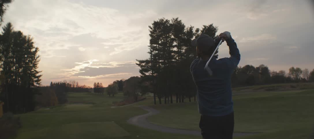 A golfer with a beard a blue shirt and a cap silhouetted against a sunset sky striking the ball on a golf course framed by distant trees