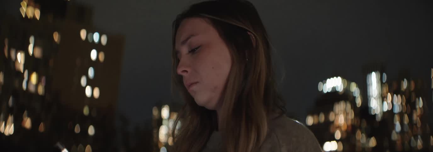 Woman with brown hair and sad expression uses a smartphone with urban illuminated buildings in the background at night