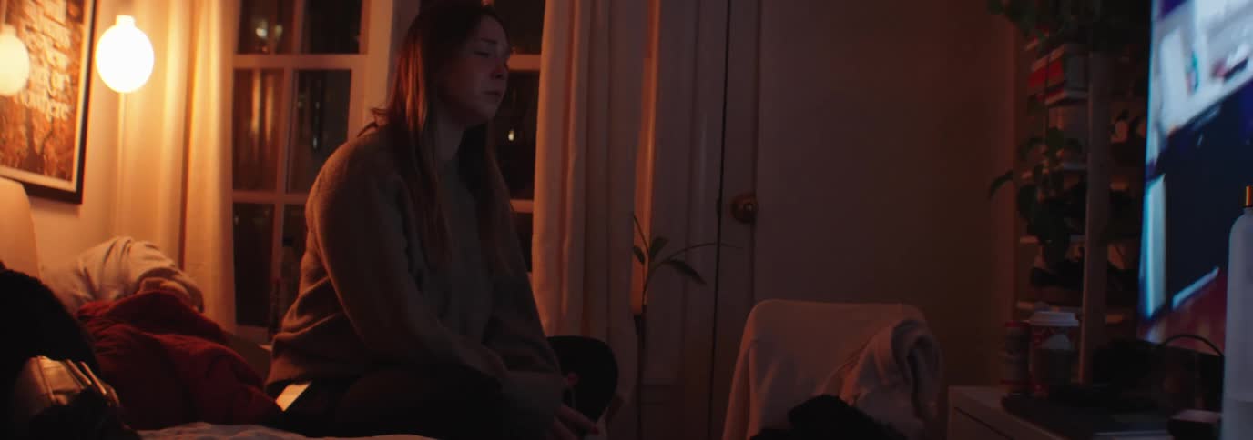 A sad woman sits on a bed in a dimly lit apartment watching television