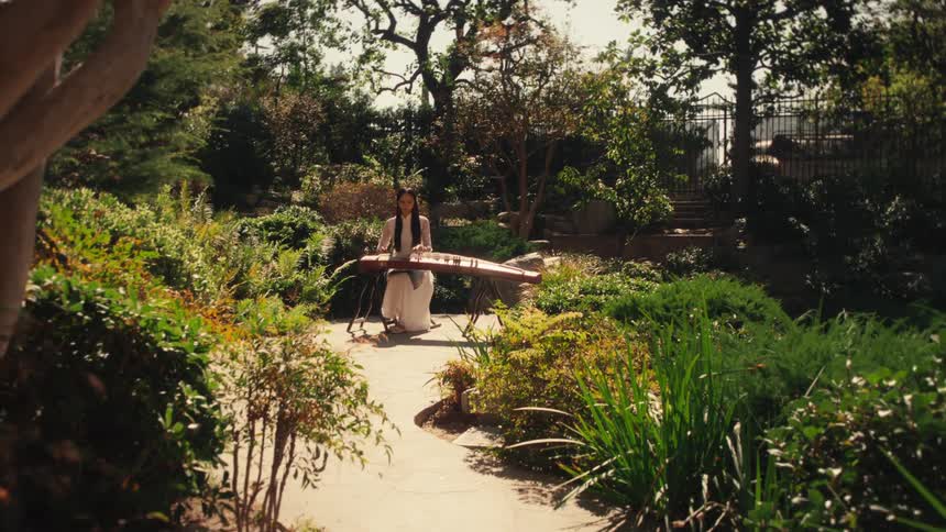 A woman in a white dress playing a guzheng a chinese zither in a lush sun dappled garden
