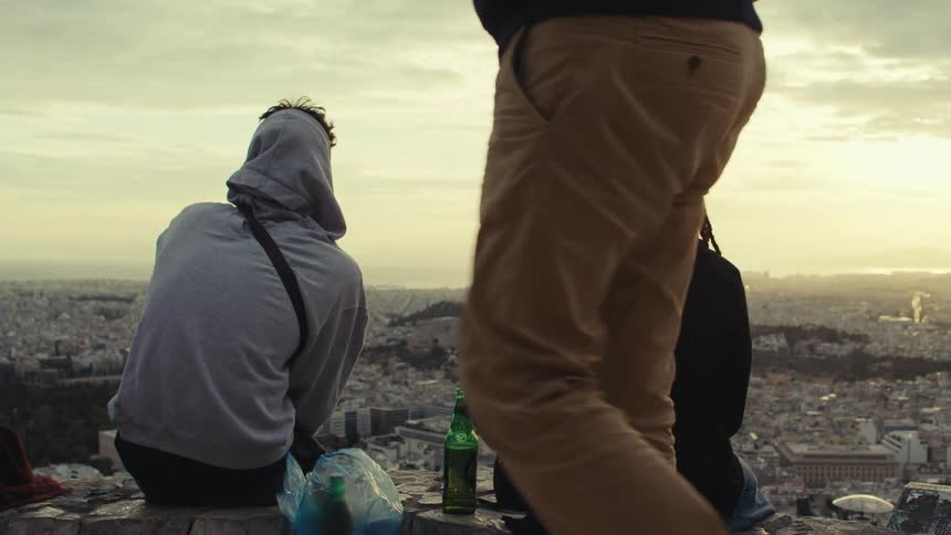 Three people standing back to back on a terrace with a panoramic view of an urban city and drinking beer