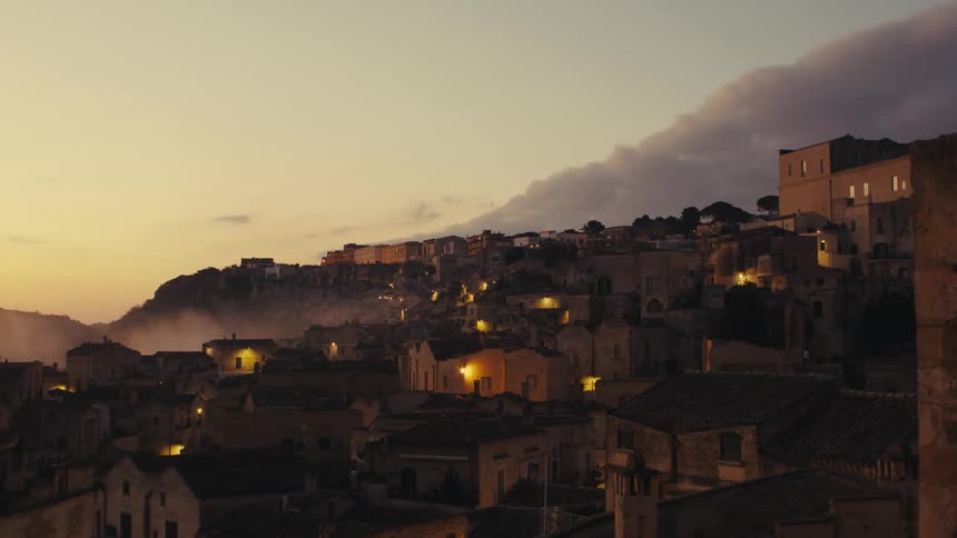 A town nestled on a hillside with a dramatic sunset casting a warm glow over the buildings and a layer of fog