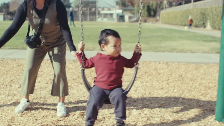 A mother gently pushes her toddler son on a swing at a playground