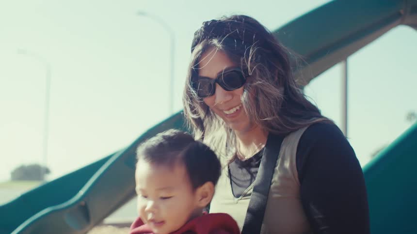 A mother and son slide down a slide in a park play area on a sunny day