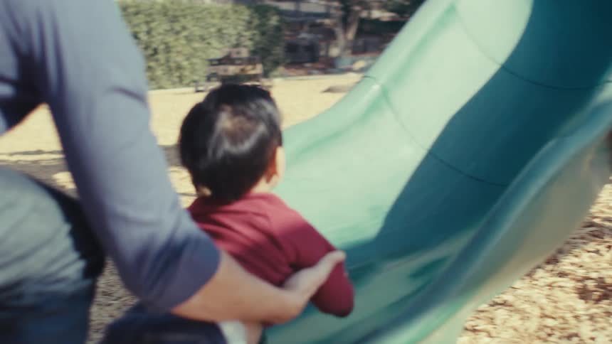 A toddler slides down a slide in a playground and his mother goes after him