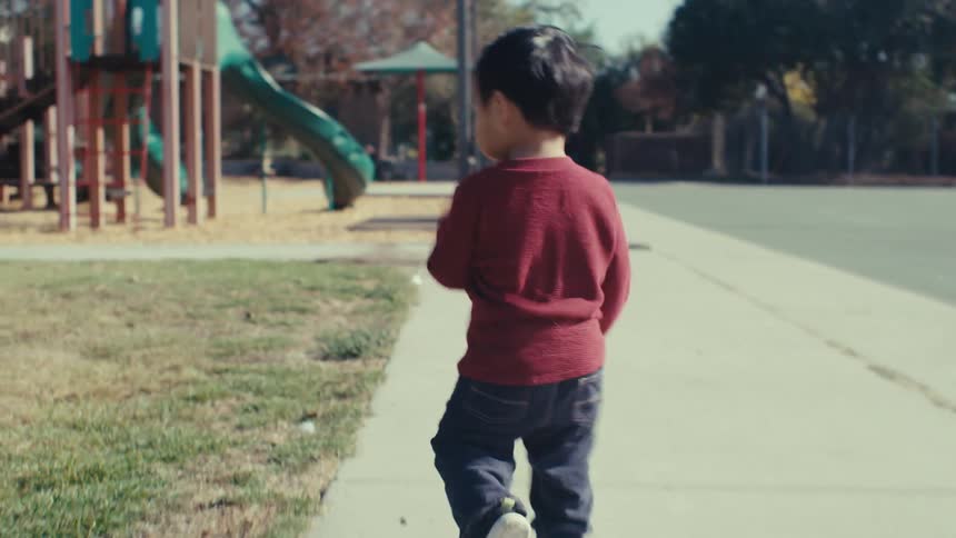 A child in a red shirt walks towards a playground in a park with trees