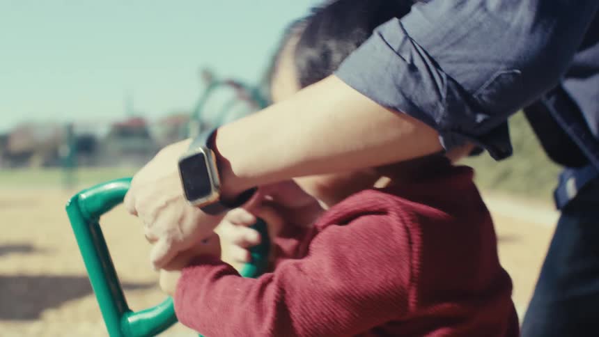 Father wearing a blue shirt and a wristwatch helps toddler son play on the playground