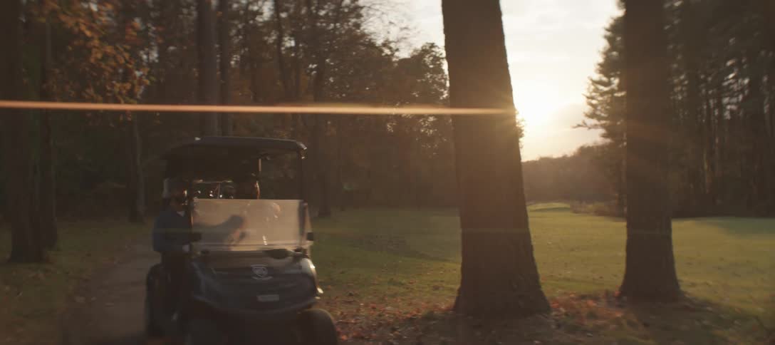 A golf cart driving through a wooded area on a golf course as the sun sets casting a warm glow across the landscape