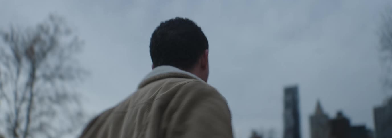 A man in a brown jacket with a white fleece collar stands impatiently under a cloudy sky in the city