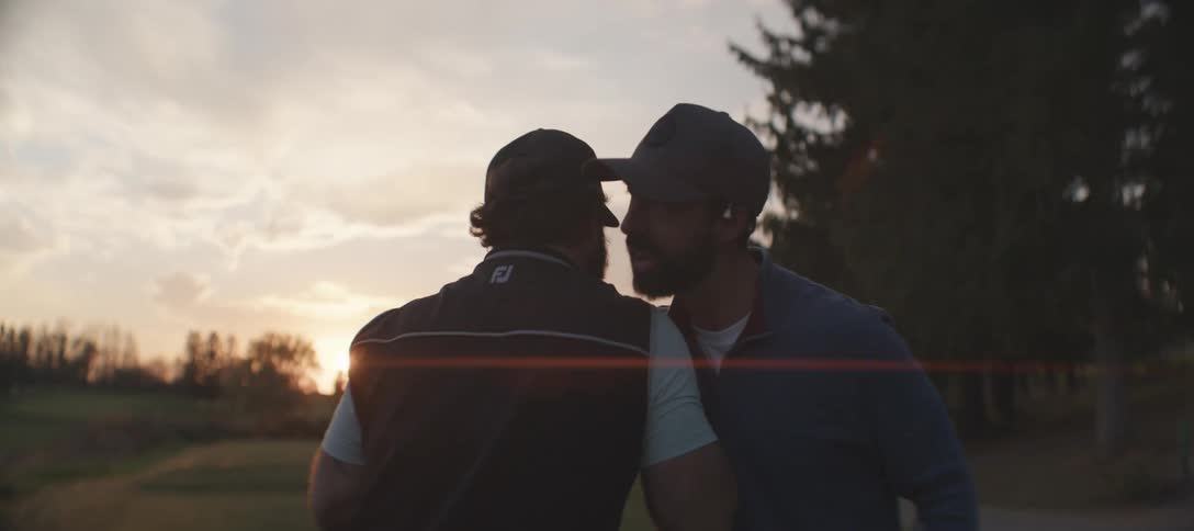 A couple of men stand on a golf course shaking hands at sunset