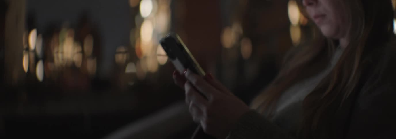 A woman with brown hair uses her smartphone against the backdrop of illuminated tall buildings at night