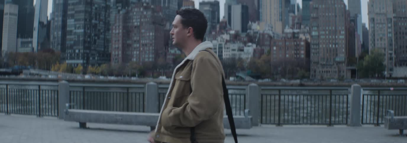 A man in a tan jacket and white shirt walking along a waterfront railing with a backdrop of modern skyscrapers and urban architecture under an overcast sky