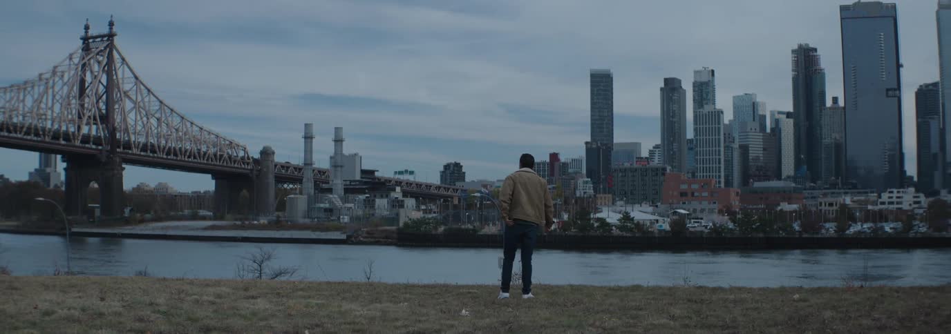 Man in a coat walking on the grass by a lake with a metropolis and a bridge in the background