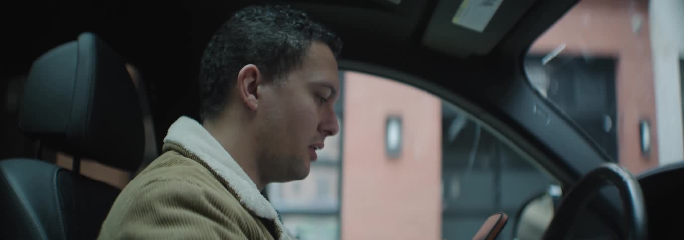 A man inside of a car reads off his smartphone and acts confused and annoyed