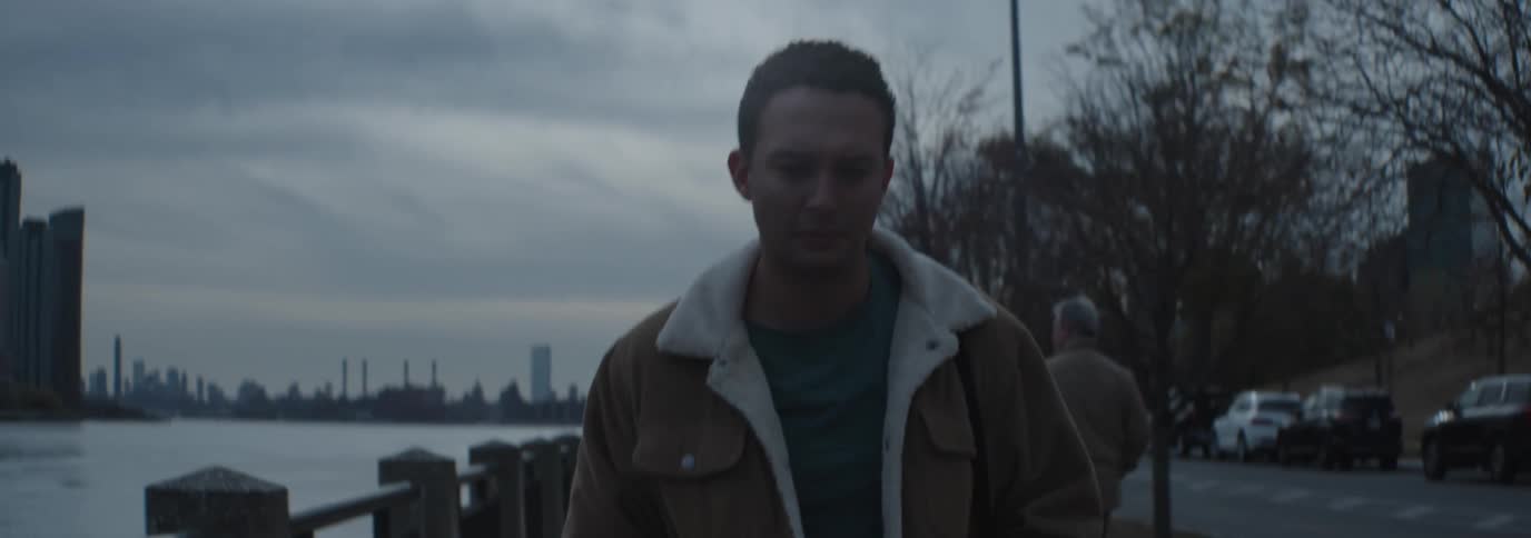 A man wearing a brown jacket with a white fleece collar walks along a riverside under a cloudy sky