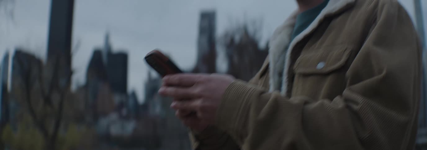 A man in a brown jacket with a white fleece collar text on his smartphone under a cloudy sky in the city