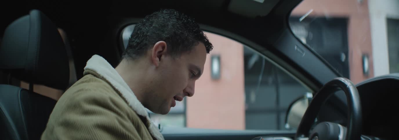 A man sits in a car in the city wearing a thick jacket moving his mouth as if reading out loud from his phone or a book