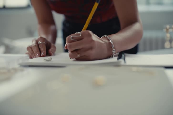 A person is writing in a notebook with a pencil wearing a bracelet and rings