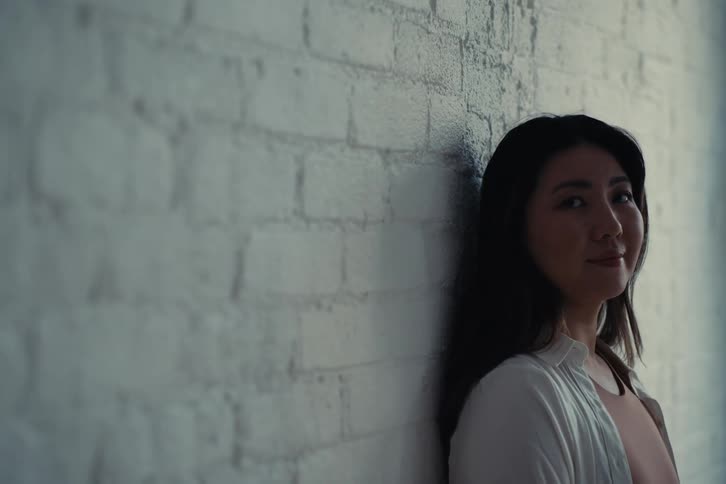 A woman with straight hair and a white shirt leans against the white brick wall smiling