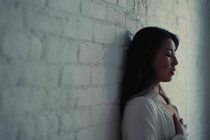 Woman with a white shirt and eyes closed leans against a white textured brick wall her hands placed on her chest