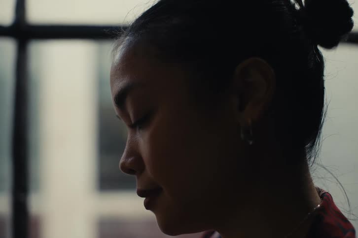 Close up of the profile of a woman with dark hair tied in a bun standing indoors near a window