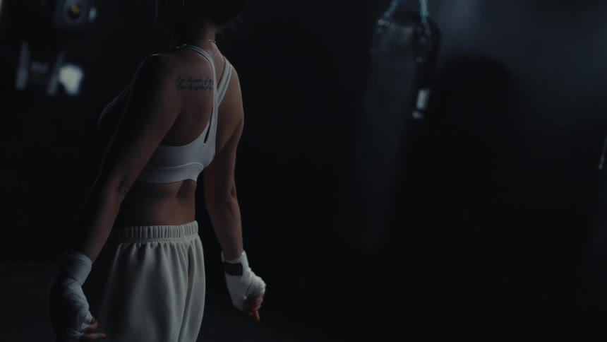 A woman in athletic wear and boxing gloves walking in the gym with a punching bag visible in the background
