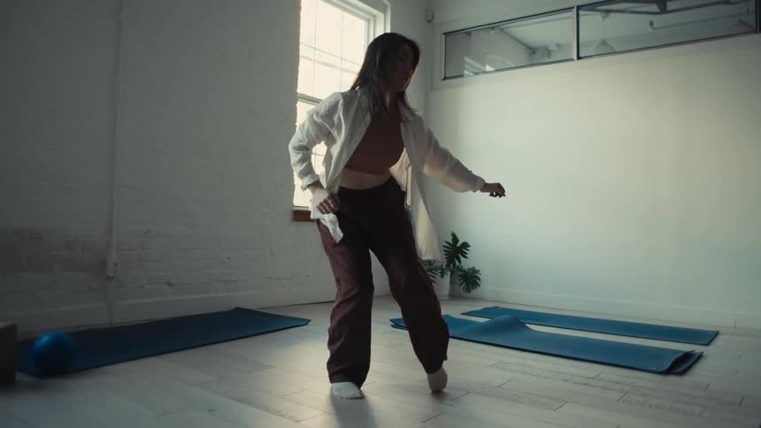 A woman dressed in loose comfortable clothing stretches in a bright minimalist room with yoga mats on the floor