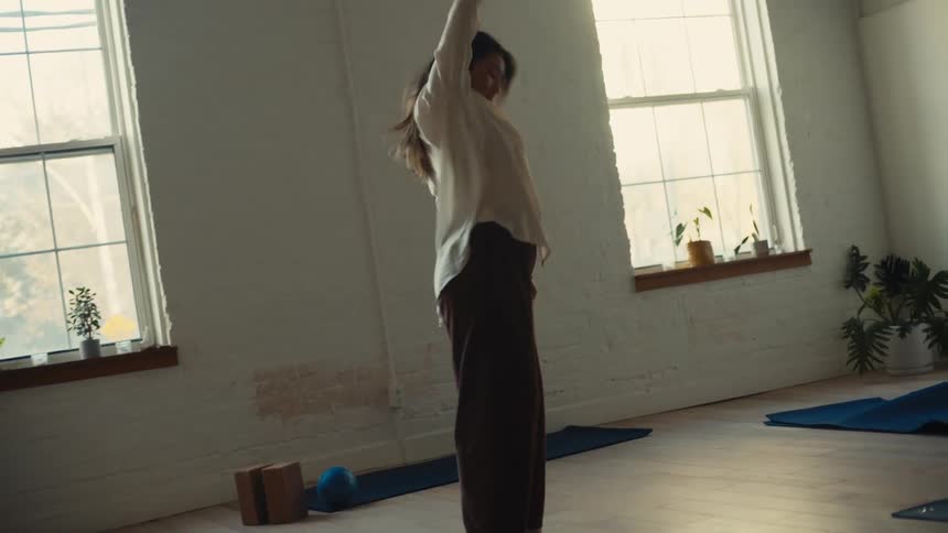 A woman practicing yoga in a spacious minimalist studio with large windows and blue yoga mats and exposed wooden beams