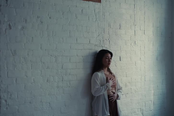 A woman with a white shirt and straight hair leans against a textured brick wall her hands placed on her chest