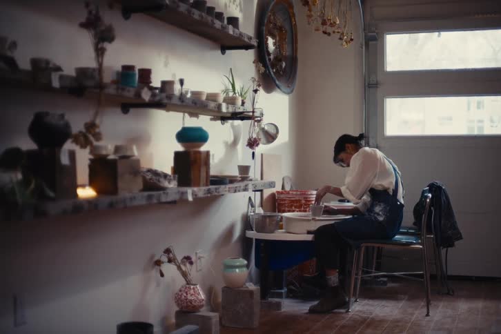A person is working on a pottery wheel in a wellorganized studio with shelves displaying various ceramic pieces and plants