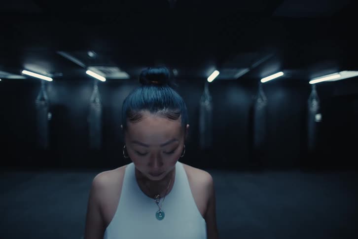 A woman in sports attire walks forward in a room with only a row of punching bags