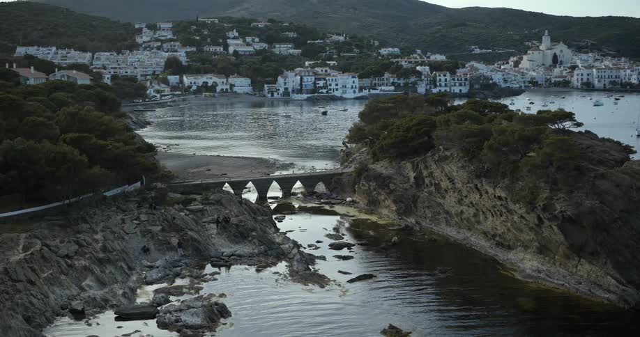 Bridge connects shore to a small islet across the water from a coastal town on a cloudy day