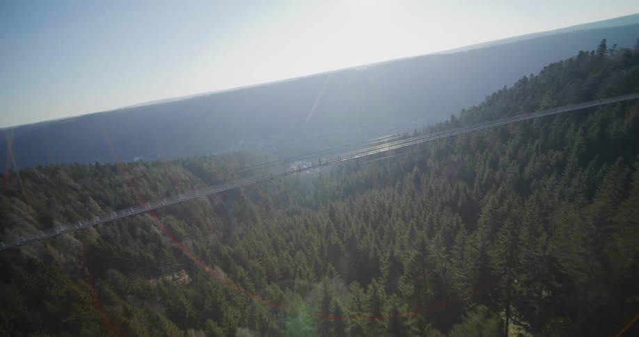 An ariel view a long suspended bridge stretching across a valley with dense green forests on either side and a clear blue sky above