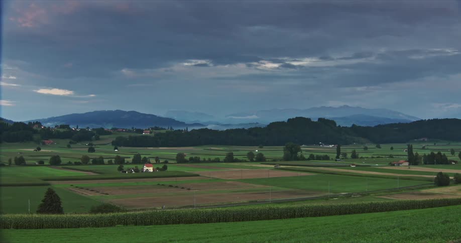 A picturesque rural landscape with a patchwork of green and brown fields scattered houses and distant hills under a cloudy sky