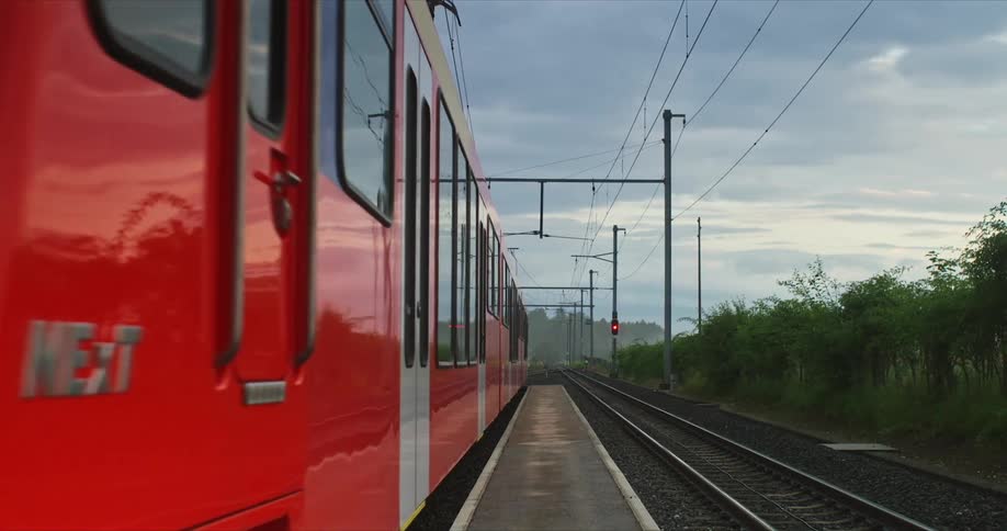 A red train is moving along a railway track under an overcast sky with power lines and poles visible above