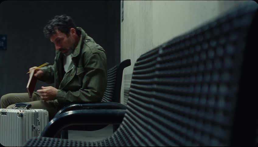 Man sitting on a bench with a silver suitcase beside him stands up