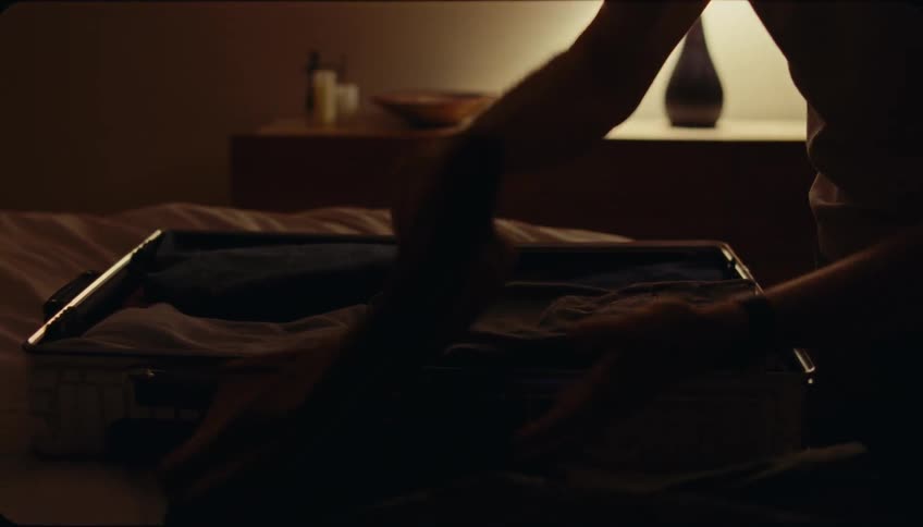Man folding clothes into a suitcase on top of a bed in a bedroom at night