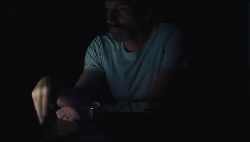 Man with a reflective and tired expression sitting in front of a laptop in a dark room