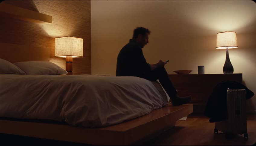 A man carries a suitcase into a hotel room at night checks his phone and then throws himself onto the bed exhausted