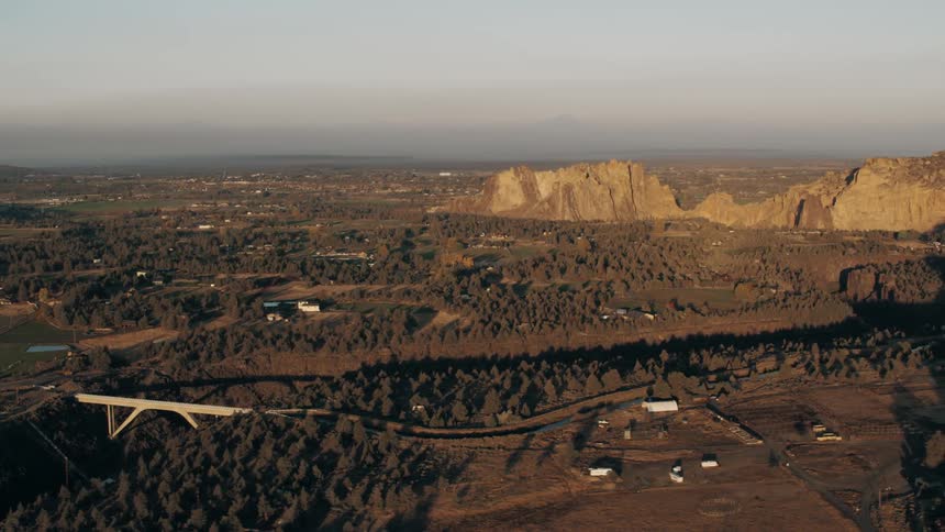 An aerial view shows a bridge crossing a river surrounded by forests and open fields with rocky cliffs and distant mountains