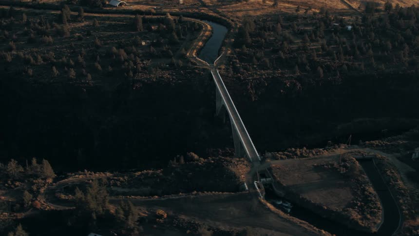 An aerial view of a bridge spanning a deep canyon with a river at the bottom surrounded by a rugged landscape with sparse vegetation and a few buildings