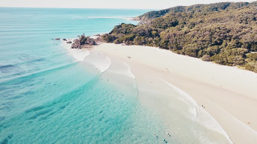 A pristine beach stretches along a coastline with clear turquoise water lapping at the shore backed by lush green hills