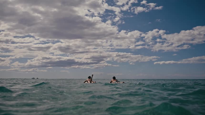 Two people are paddling on surfboards in a calm ocean under a partly cloudy sky