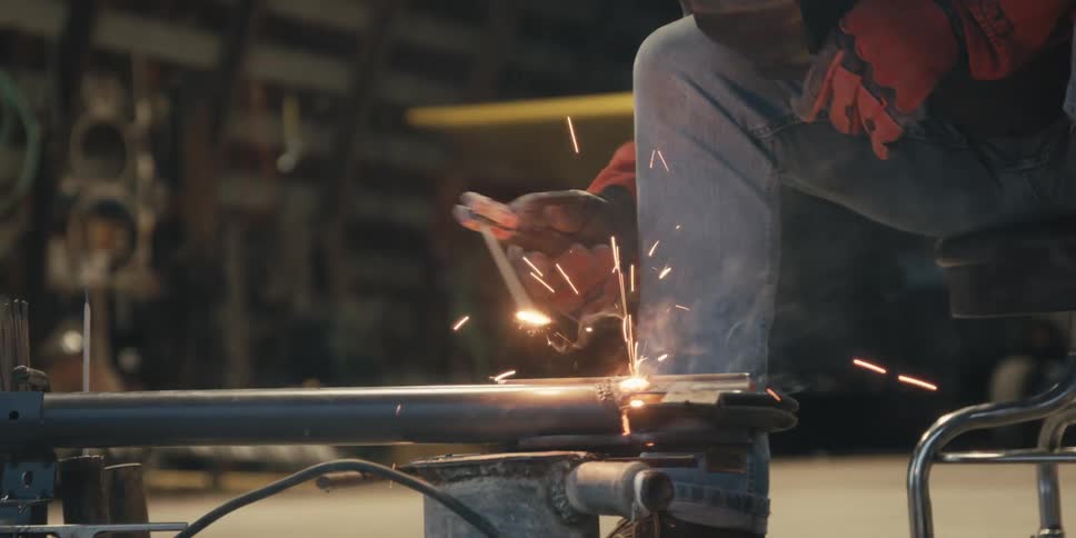 Male worker wearing protective gear sitting down on a stool indoors soldering a piece of metal